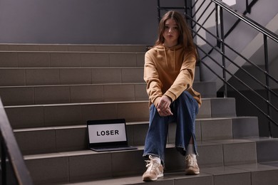 Photo of Cyber bullying. Upset teenage girl and laptop with text You Loser on stairs indoors. Space for text