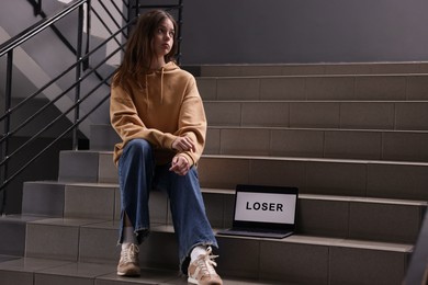 Photo of Cyber bullying. Upset teenage girl and laptop with text You Loser on stairs indoors. Space for text