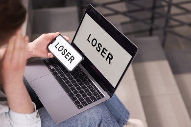 Photo of Cyber bullying. Upset teenage girl looking at laptop and smartphone with text You Loser on stairs indoors, closeup