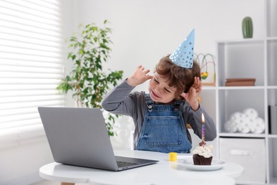Photo of Little boy celebrating his birthday while having video call via laptop at table indoors
