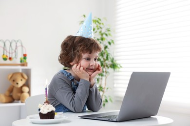 Photo of Birthday celebration. Little boy with cupcake having video call via laptop at table indoors