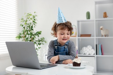 Photo of Birthday celebration. Little boy with cupcake having video call via laptop at table indoors