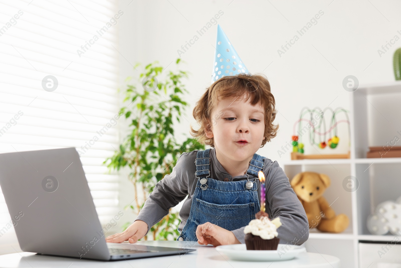 Photo of Birthday celebration. Little boy blowing candle on cupcake while having video call via laptop at table indoors