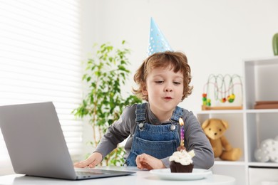Photo of Birthday celebration. Little boy blowing candle on cupcake while having video call via laptop at table indoors