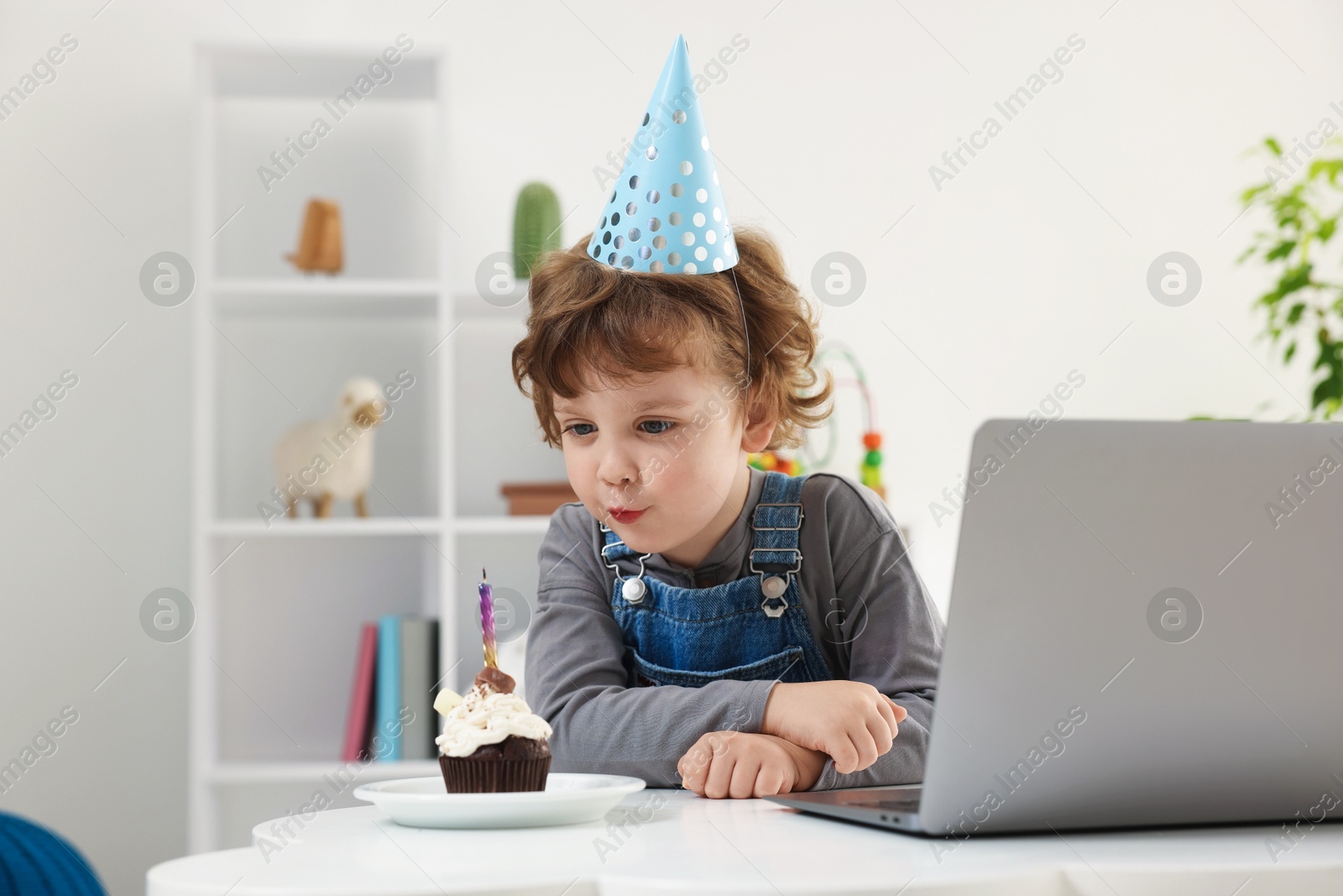 Photo of Birthday celebration. Little boy blowing candle on cupcake while having video call via laptop at table indoors