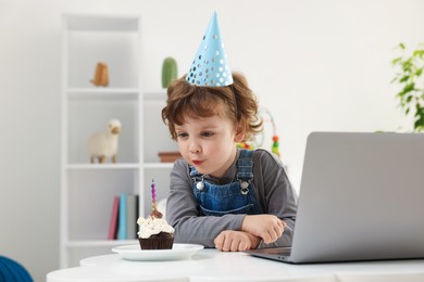 Photo of Birthday celebration. Little boy blowing candle on cupcake while having video call via laptop at table indoors