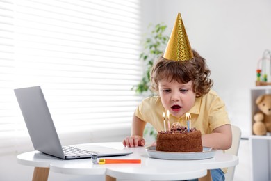 Photo of Birthday celebration. Little boy blowing candles on cake while having video call via laptop at table indoors. Space for text
