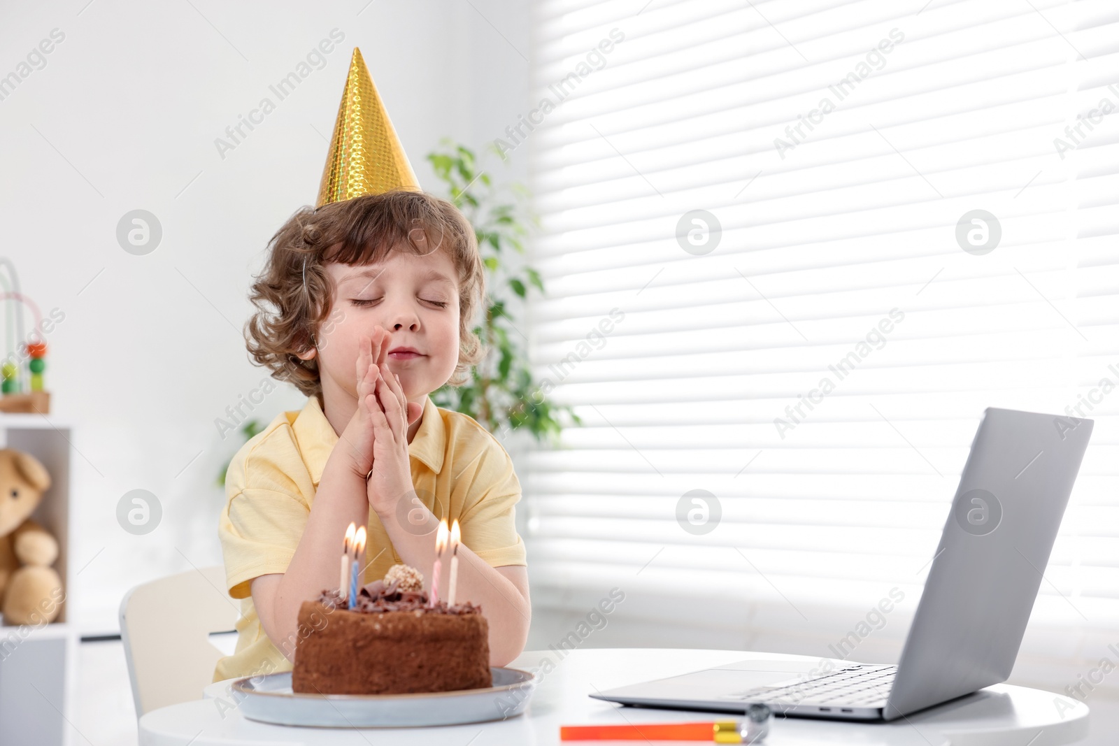 Photo of Birthday celebration. Little boy with cake making wish while having video call via laptop at table indoors