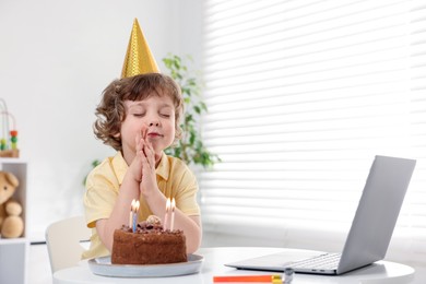 Photo of Birthday celebration. Little boy with cake making wish while having video call via laptop at table indoors