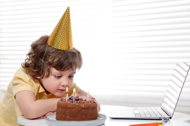 Photo of Birthday celebration. Little boy with cake having video call via laptop at table indoors. Space for text