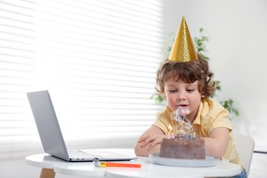 Photo of Birthday celebration. Little boy with cake having video call via laptop at table indoors. Space for text