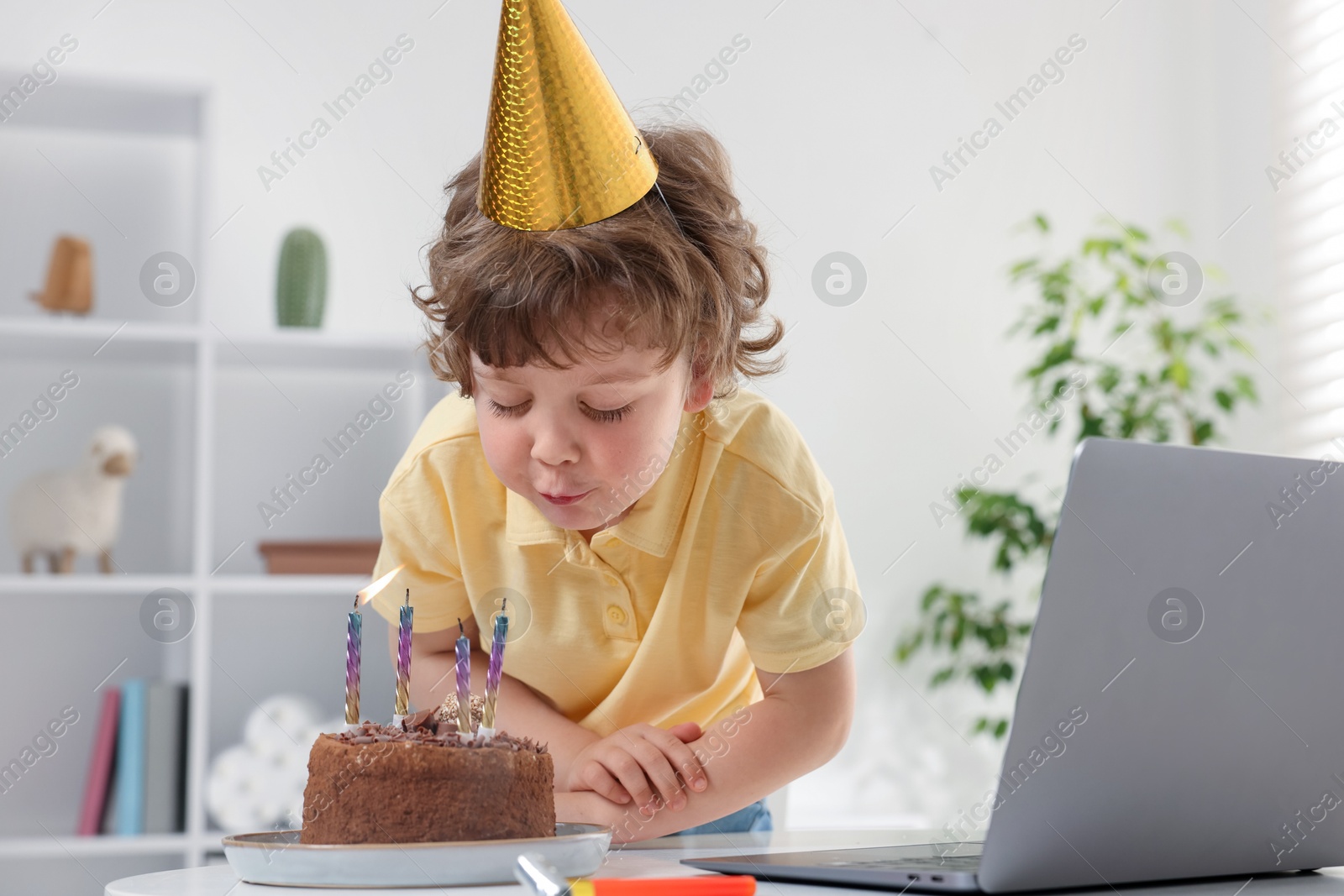 Photo of Birthday celebration. Little boy blowing candles on cake while having video call via laptop at table indoors