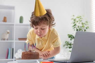 Photo of Birthday celebration. Little boy blowing candles on cake while having video call via laptop at table indoors