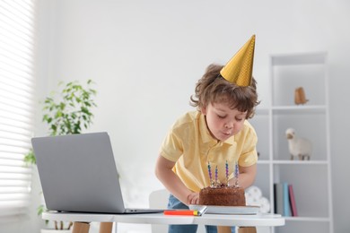 Photo of Birthday celebration. Little boy blowing candles on cake while having video call via laptop at table indoors