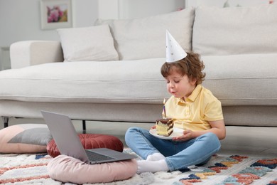 Photo of Birthday celebration. Little boy blowing candle on piece of cake while having video call via laptop at home