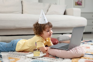 Photo of Birthday celebration. Little boy with piece of cake having video call via laptop at home