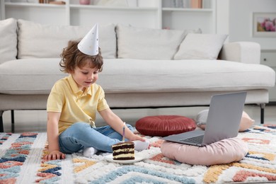 Photo of Birthday celebration. Little boy with piece of cake having video call via laptop at home