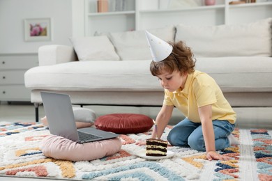 Photo of Birthday celebration. Little boy blowing candle on piece of cake while having video call via laptop at home