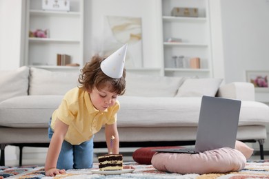 Photo of Birthday celebration. Little boy blowing candle on piece of cake while having video call via laptop at home