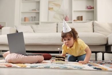 Photo of Birthday celebration. Little boy blowing candle on piece of cake while having video call via laptop at home