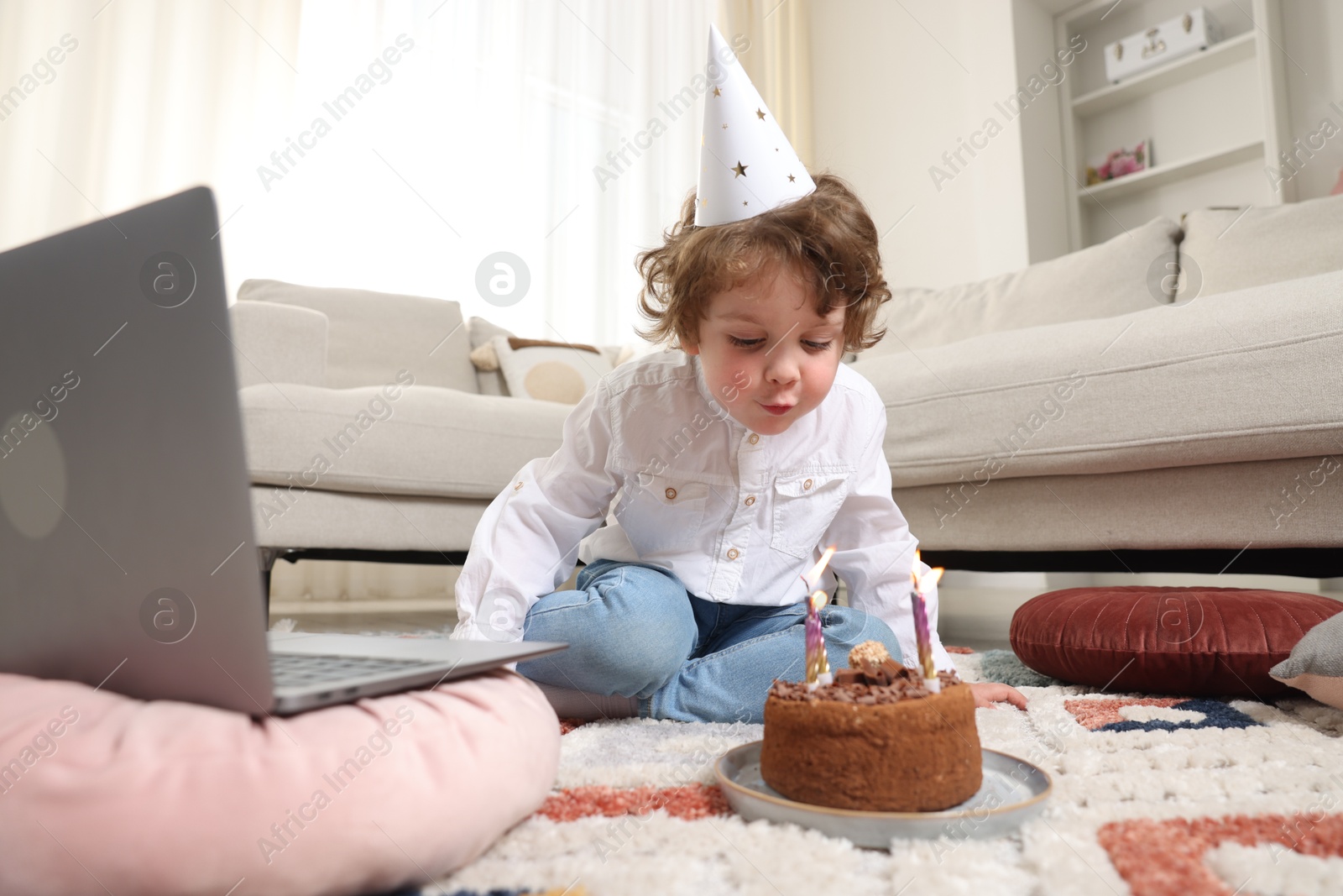 Photo of Birthday celebration. Little boy blowing candles on cake while having video call via laptop at home