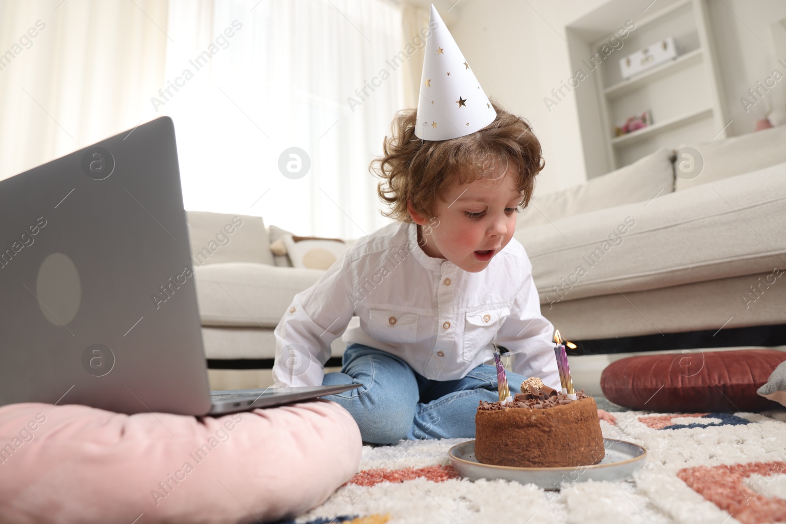 Photo of Birthday celebration. Little boy blowing candles on cake while having video call via laptop at home