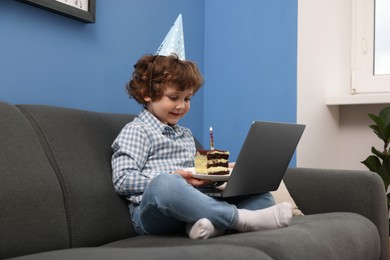 Photo of Birthday celebration. Little boy with piece of cake having video call via laptop on sofa at home