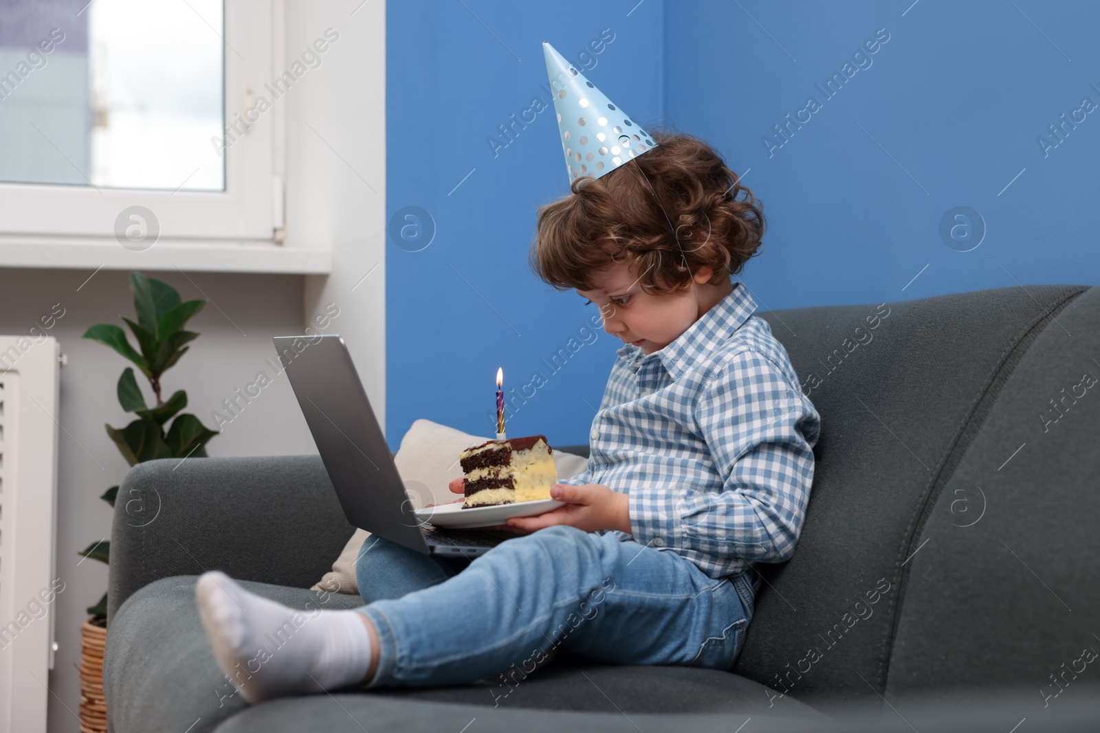 Photo of Birthday celebration. Little boy with piece of cake having video call via laptop on sofa at home