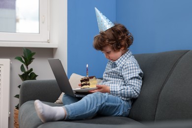 Photo of Birthday celebration. Little boy with piece of cake having video call via laptop on sofa at home