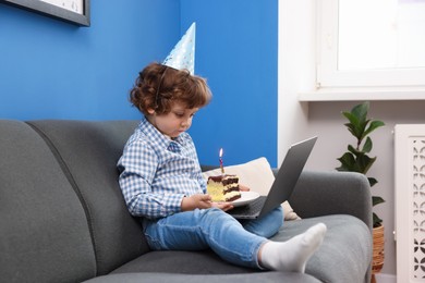 Photo of Birthday celebration. Little boy with piece of cake having video call via laptop on sofa at home