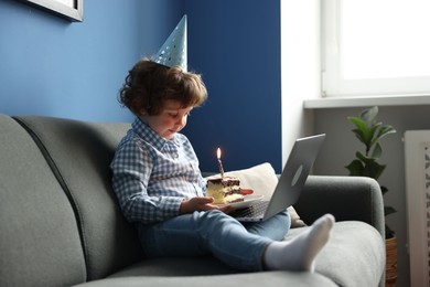 Photo of Birthday celebration. Little boy with piece of cake having video call via laptop on sofa at home