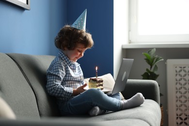 Photo of Birthday celebration. Little boy with piece of cake having video call via laptop on sofa at home