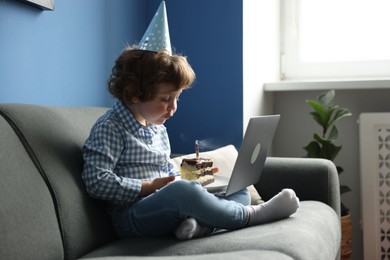 Photo of Birthday celebration. Little boy blowing candle on piece of cake while having video call via laptop on sofa at home