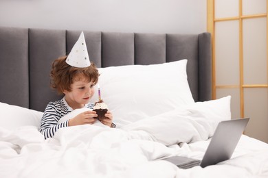 Photo of Birthday celebration. Little boy with cupcake having video call via laptop in bed at home