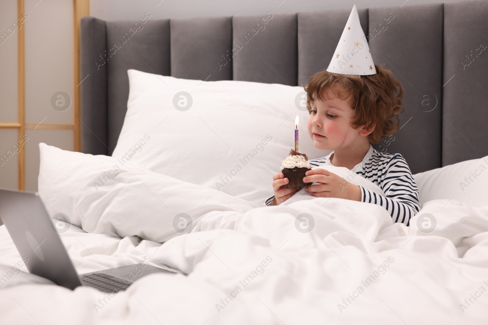 Photo of Birthday celebration. Little boy with cupcake having video call via laptop in bed at home