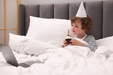 Photo of Birthday celebration. Little boy with cupcake having video call via laptop in bed at home