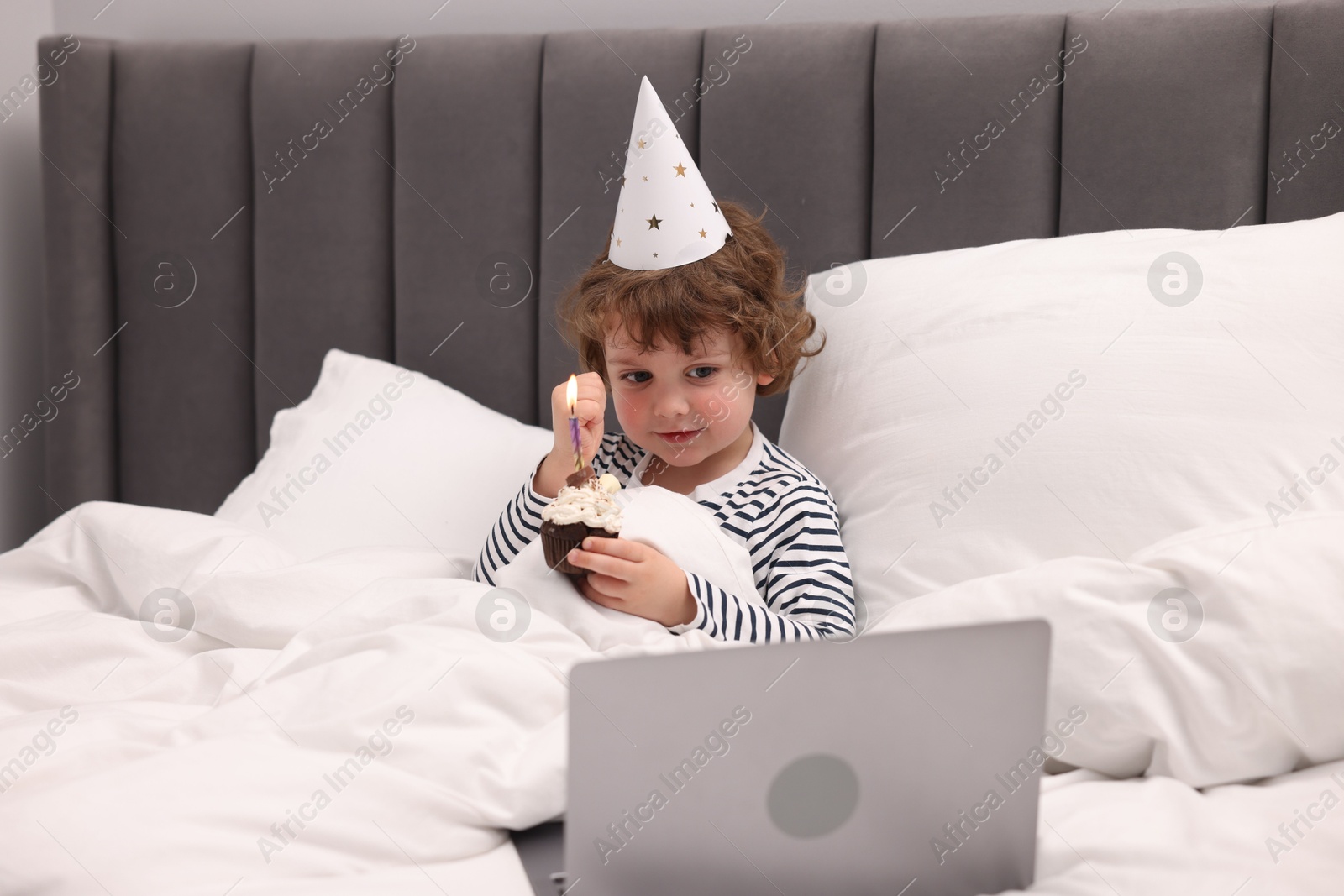 Photo of Birthday celebration. Little boy with cupcake having video call via laptop in bed at home