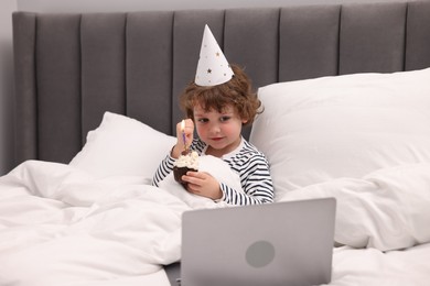 Photo of Birthday celebration. Little boy with cupcake having video call via laptop in bed at home