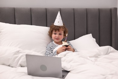 Photo of Birthday celebration. Little boy with cupcake having video call via laptop in bed at home