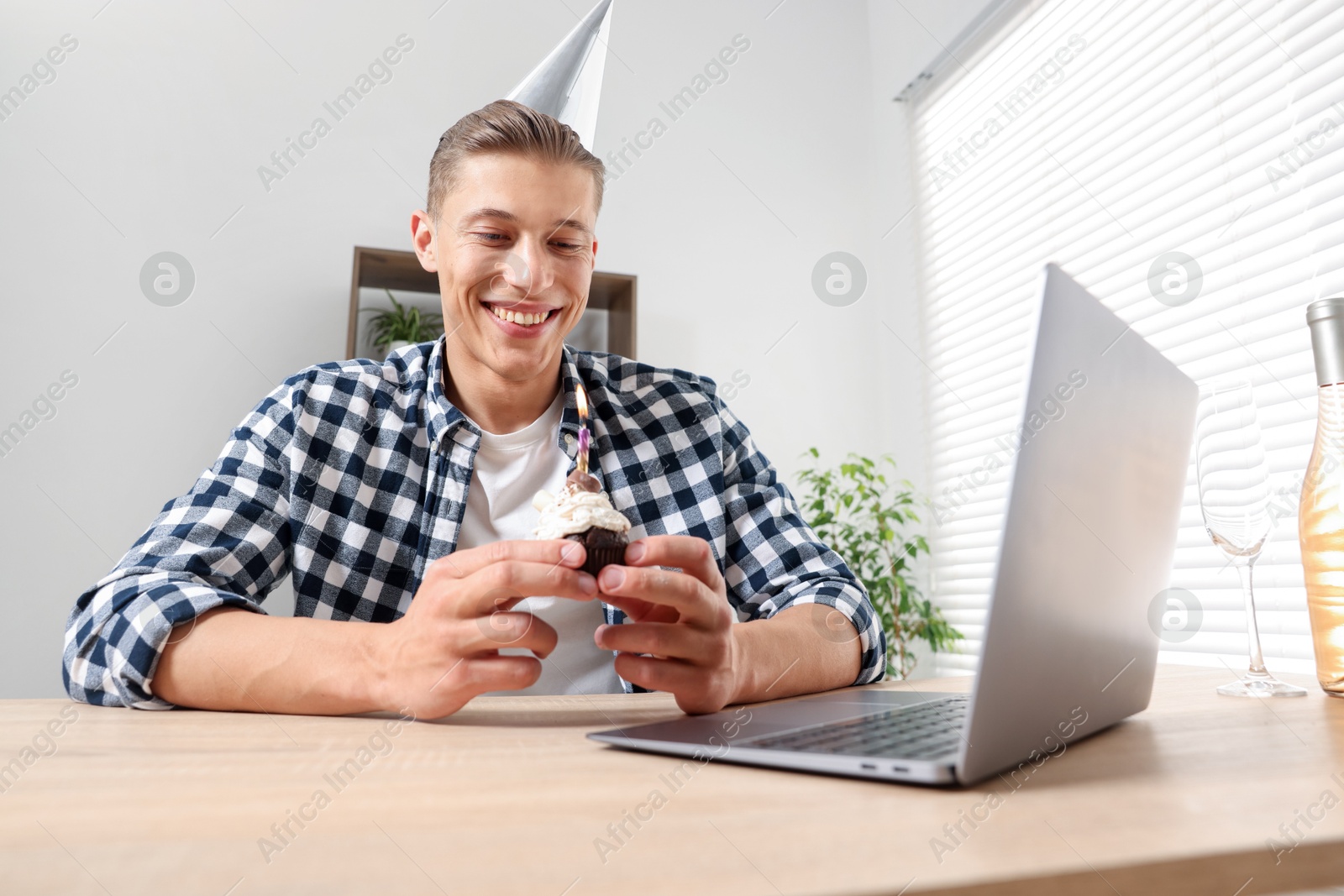 Photo of Birthday celebration. Man with cupcake having video call via laptop at table indoors, low angle view