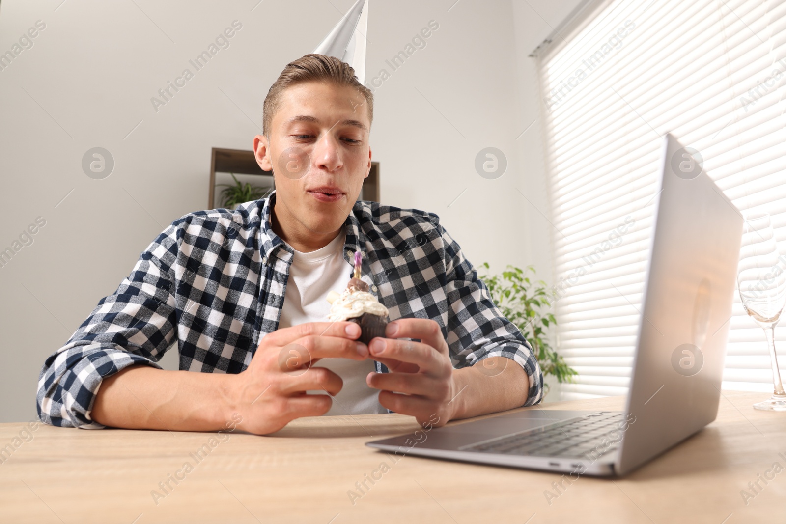 Photo of Birthday celebration. Man blowing candle on cupcake while having video call via laptop at desk indoors, low angle view