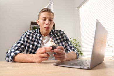 Photo of Birthday celebration. Man blowing candle on cupcake while having video call via laptop at desk indoors, low angle view