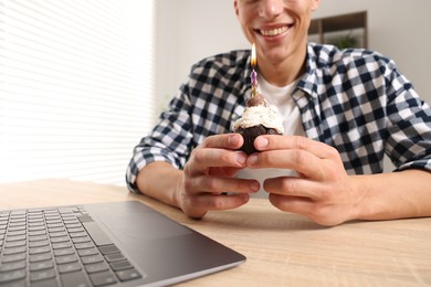 Photo of Birthday celebration. Man with cupcake having video call via laptop at table indoors, closeup