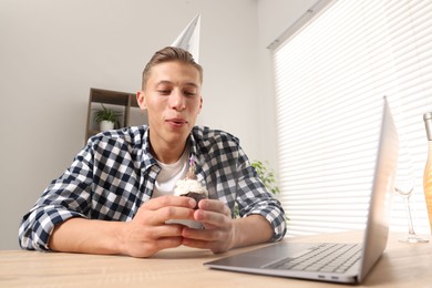 Photo of Birthday celebration. Man blowing candle on cupcake while having video call via laptop at desk indoors, low angle view