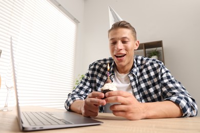 Photo of Birthday celebration. Man blowing candle on cupcake while having video call via laptop at desk indoors, low angle view