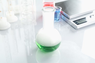 Photo of Flask with green liquid, other laboratory glassware and scale on white mirror table, closeup