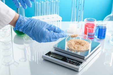 Photo of Scientist weighing Petri dish with sample on scales at white mirror table against light blue background, closeup