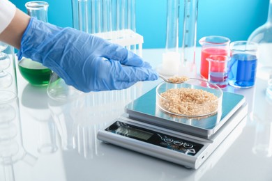 Photo of Scientist weighing Petri dish with sample on scales at white mirror table against light blue background, closeup