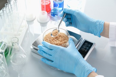 Photo of Scientist weighing Petri dish with sample on scales at white mirror table, closeup