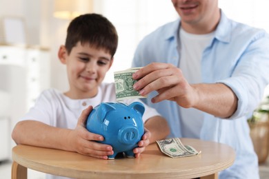 Photo of Father and son putting pocket money into piggybank at table indoors, selective focus